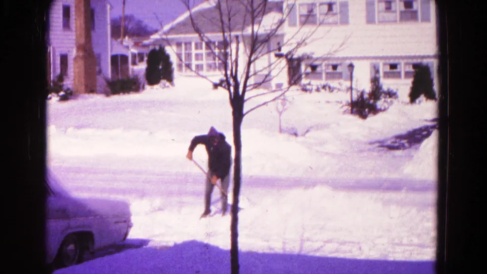 A man in a dark hooded jacket shovels snow from a driveway area in a snowy Hagerstown neighborhood.