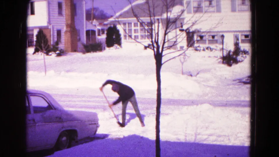 1960s home-movie view of a man lifting snow near a driveway, with a sedan, houses, and a bare tree in frame.