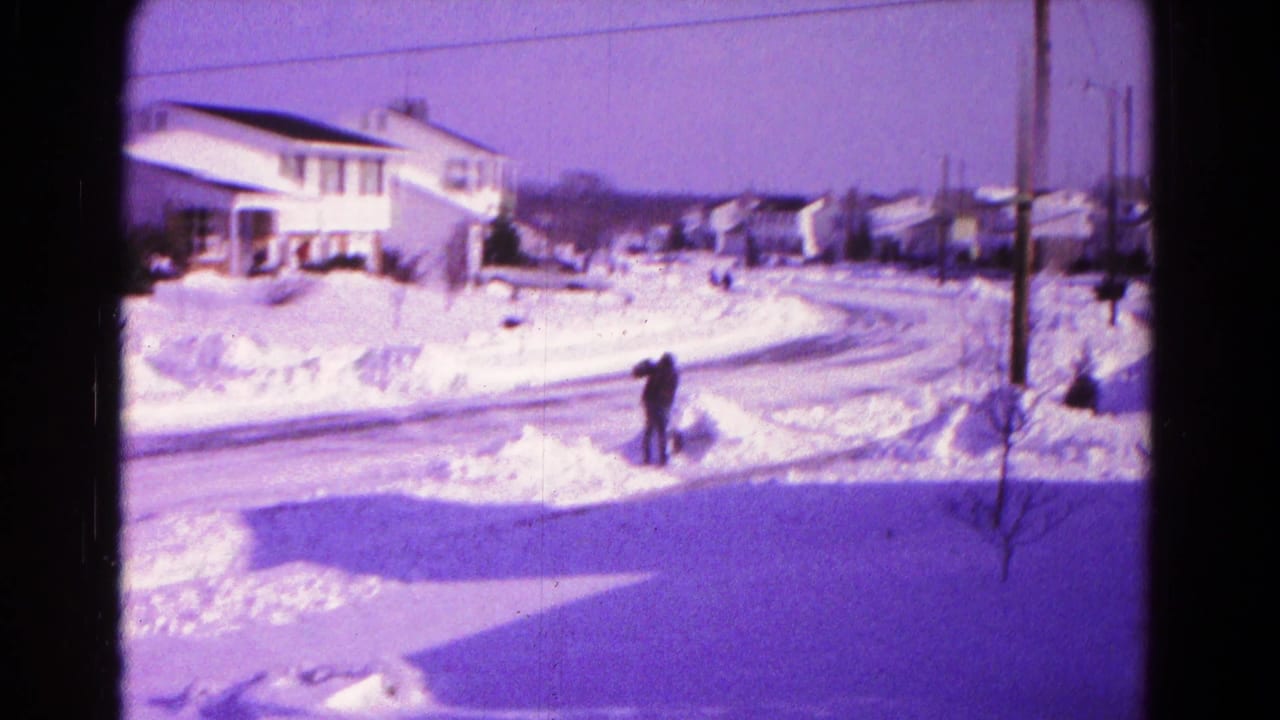 Snow-covered suburban street with a man shoveling near piled snowbanks.