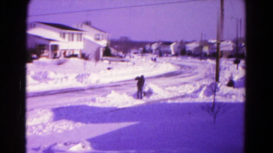 1966 Hagerstown home-movie frame showing a man shoveling snow beside a residential street lined with houses.