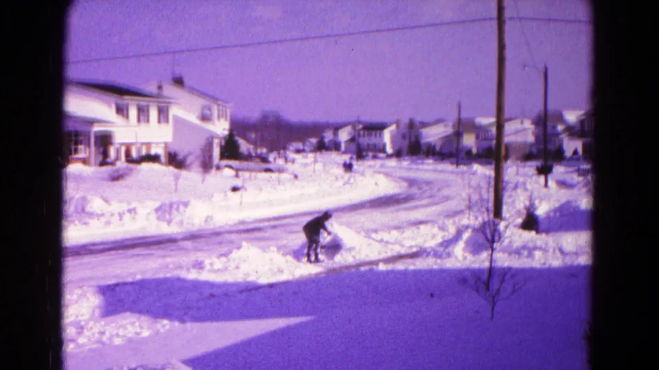 A man bends into the work of shoveling snow beside a Hagerstown street, with snowbanks and mid-century homes behind him.