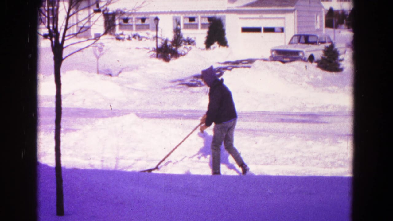 Man in a dark coat and hat shovels snow in a Hagerstown, Maryland driveway during the 1966 winter scene.