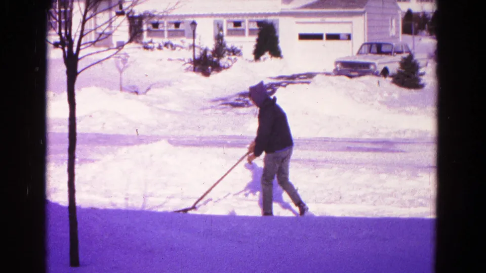 Man in a dark coat and hat shovels snow in a Hagerstown, Maryland driveway during the 1966 winter scene.