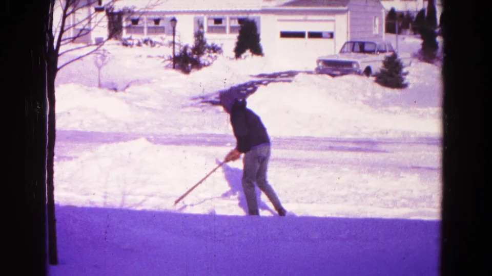 Adult man continues shoveling snow outside a suburban home, with a parked car and snowbanks in the background.