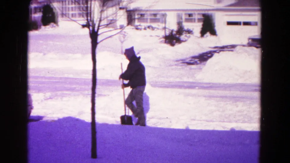 Man pauses with a shovel in the snowy residential driveway scene, framed by trees, snowbanks, and a suburban house.