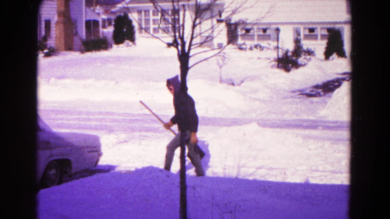 Bundled person carrying a snow shovel beside a parked sedan on a snowy suburban street.