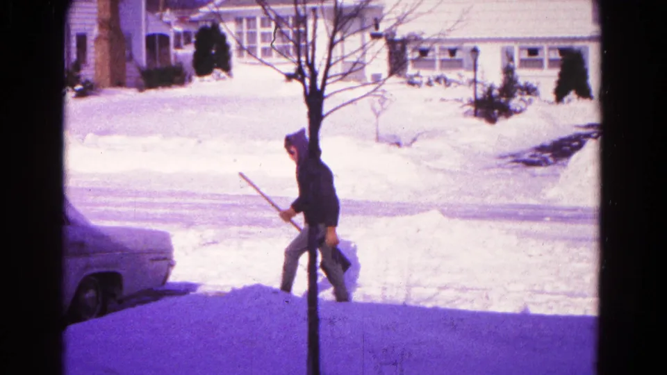 In 1966 Hagerstown, Maryland, a bundled young person carries a snow shovel near a parked sedan with snowbanks and houses behind them.