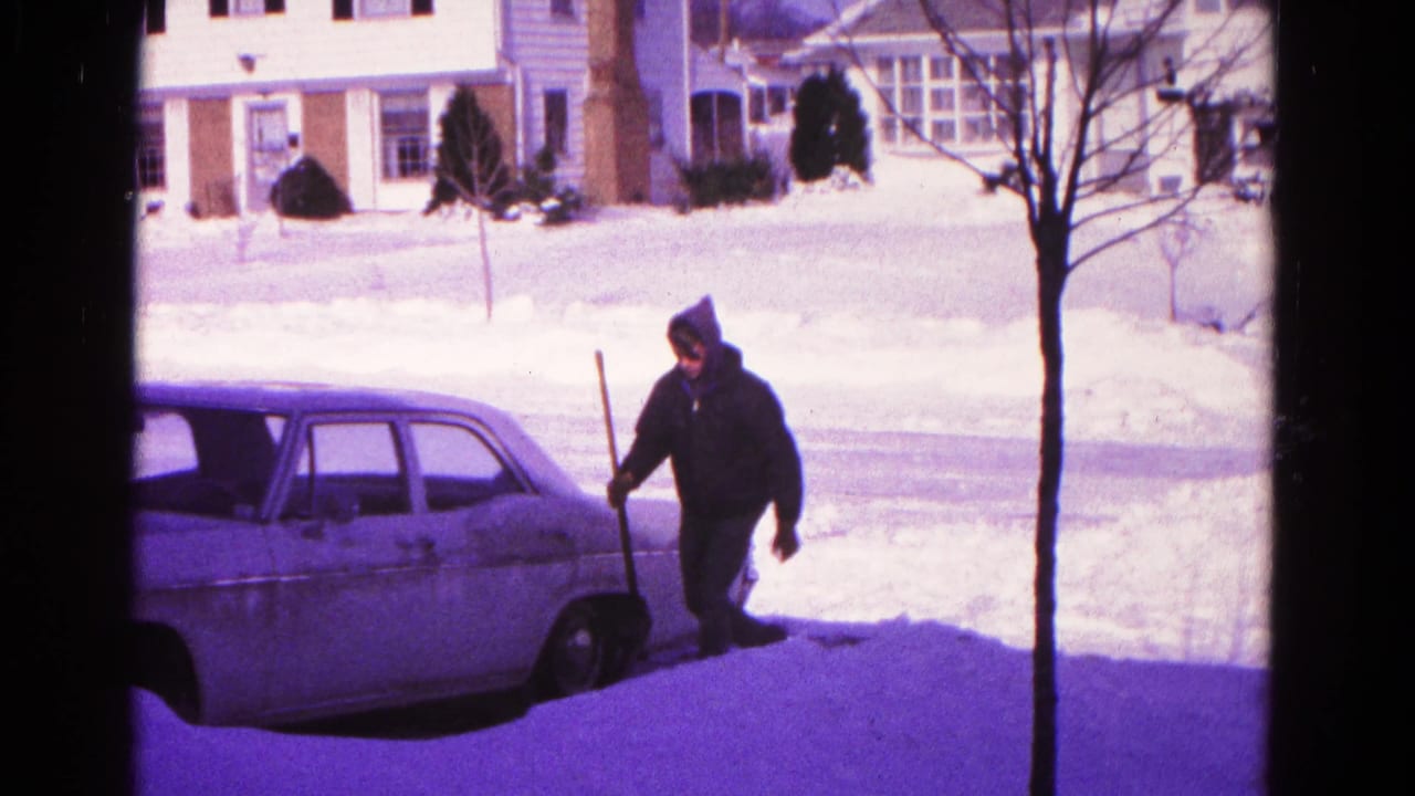 Person with a snow shovel walks near the front of a parked car along a snowy curb.