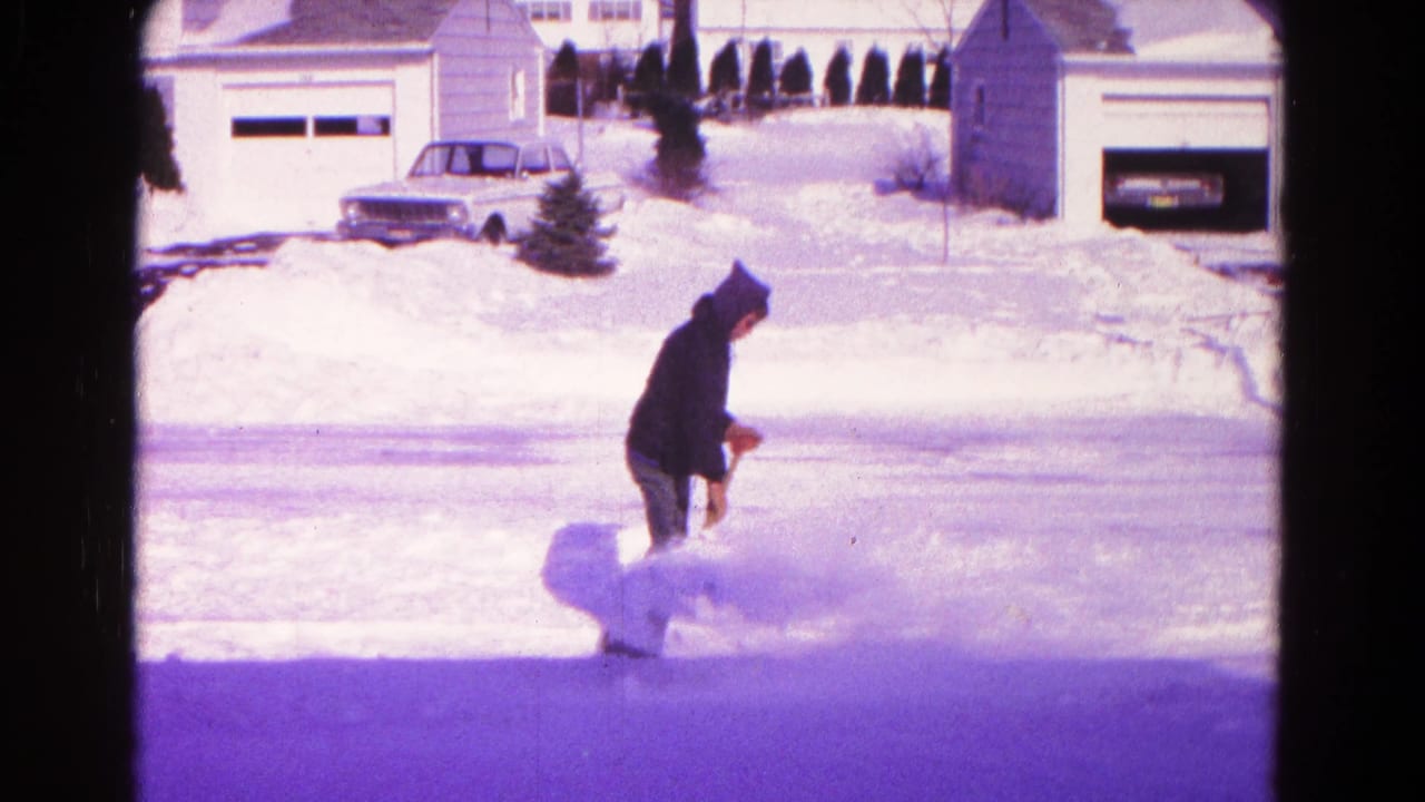 In 1966 Hagerstown home-movie footage, a hooded man starts clearing snow beside a suburban street with garages and a parked car in the background.