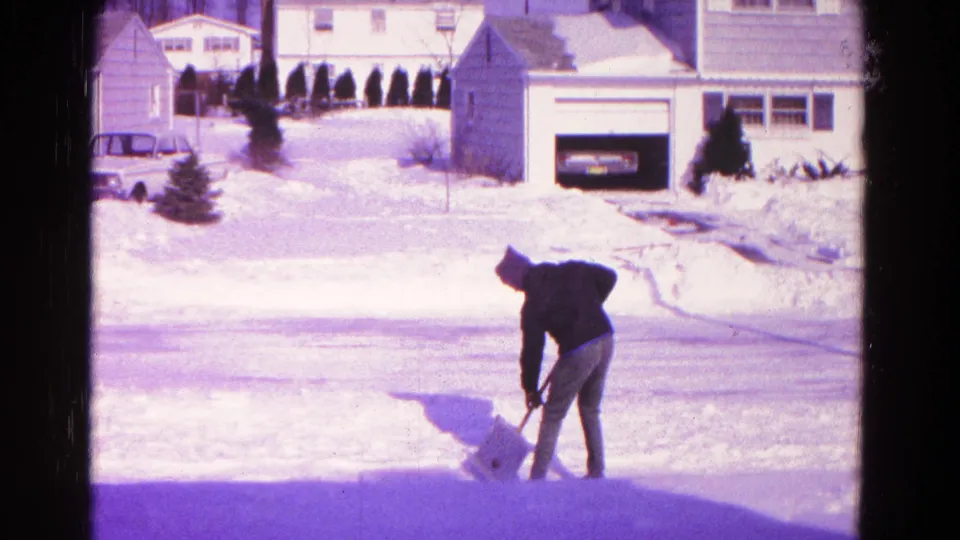 The man bends to scoop snow along a suburban roadside, framed by 1960s home-movie color and snow-covered houses behind him.