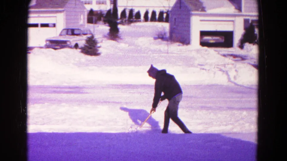A hooded man continues clearing snow at the street edge in the 1966 Hagerstown clip, with a parked sedan and garages visible across the road.