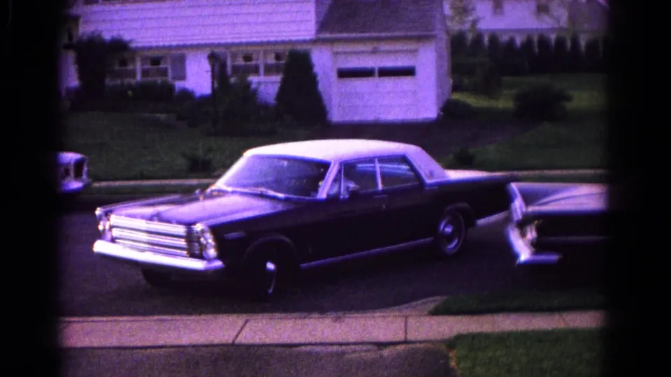 The sedan continues backing or turning onto the street, with a white house, garage, and lawn visible behind it.
