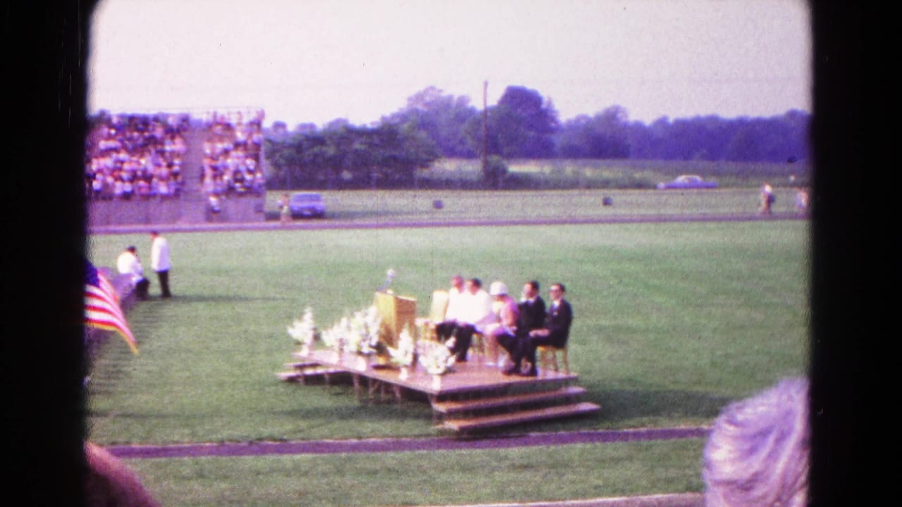 Outdoor graduation platform on a grass field with seated people, podium, flowers, bleachers, and an American flag.