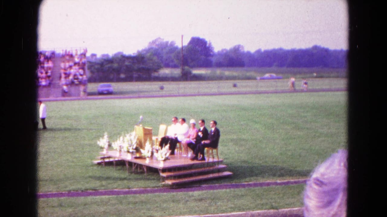 Small graduation stage on a field with people seated beside a podium and spectators in bleachers behind the track.