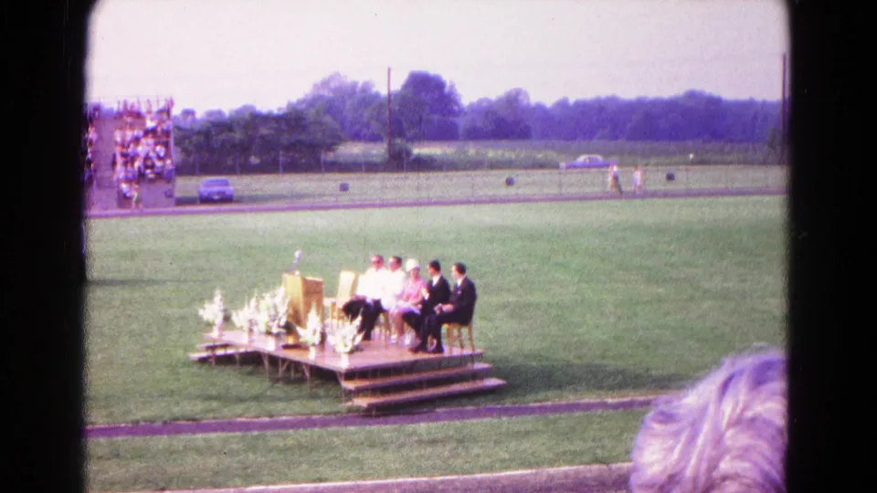 A spectator view of the Hagerstown graduation field shows the stage, seated officials or graduates, flowers, bleachers, track, and distant cars.