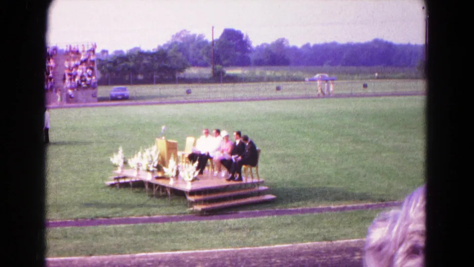 1966 Hagerstown outdoor commencement scene with a platform on the field, seated participants, podium, floral decor, bleachers, and spectator foreground.