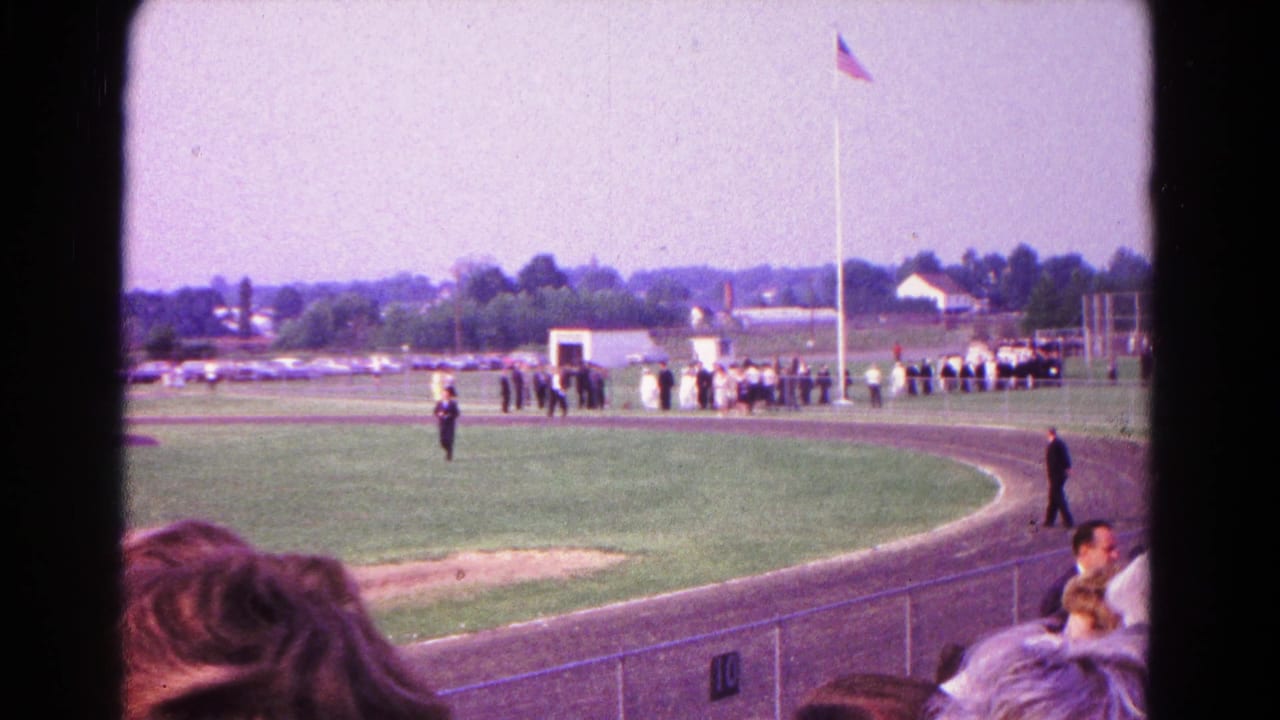 1966 Hagerstown home-movie frame showing a racetrack, grassy infield, spectators, and people lined near a U.S. flagpole.