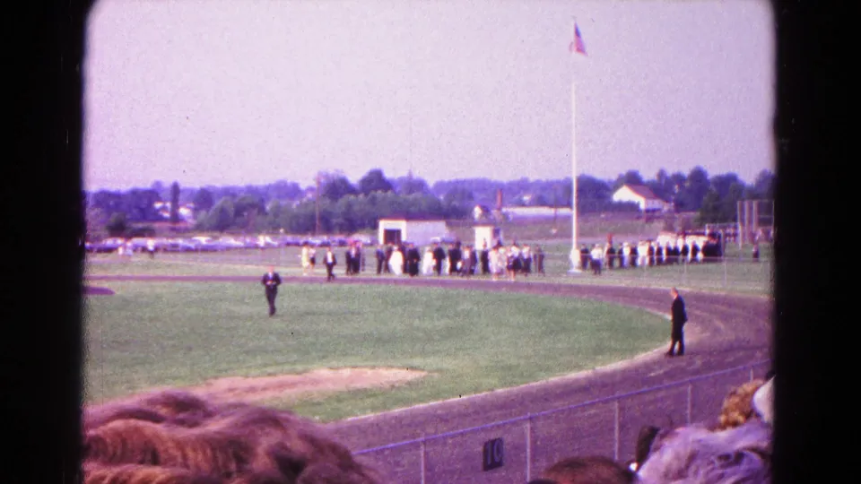 Wide 1966 Hagerstown frame from the stands, with a racetrack, infield, distant buildings, and a group assembled near a U.S. flag.