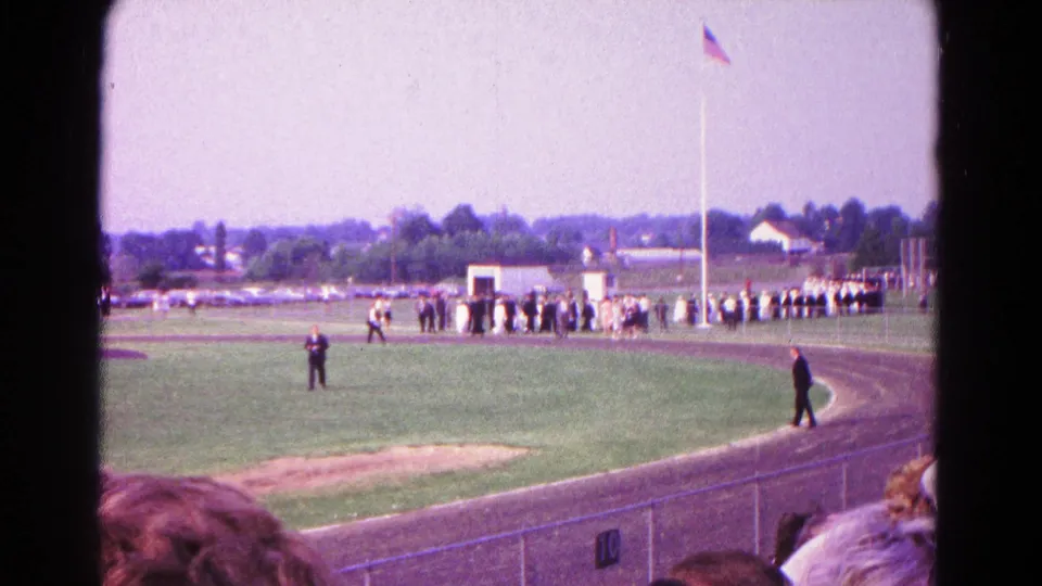 1966 Hagerstown home-movie still showing people gathered by the flagpole, a curved track, spectators in front, and open fields beyond.