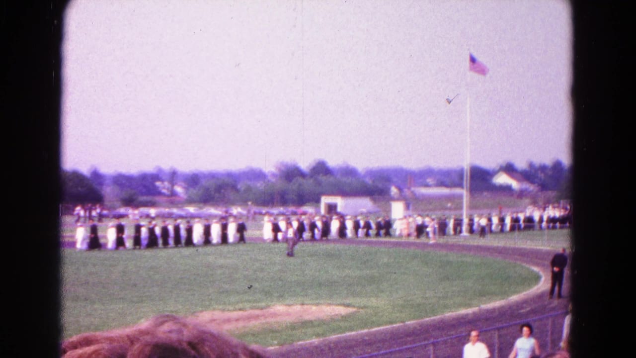 1966 Hagerstown home-movie view of a formal line on a track field, with spectators in the foreground and an American flag overhead.