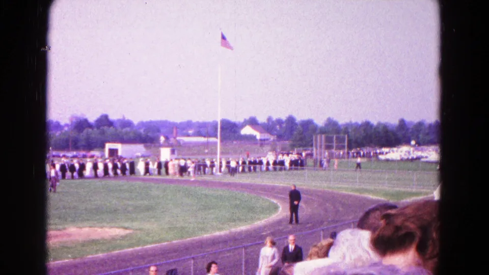 Home-movie frame from Hagerstown in 1966 showing a ceremony line across the field, with bleachers, spectators, and a flagpole visible.