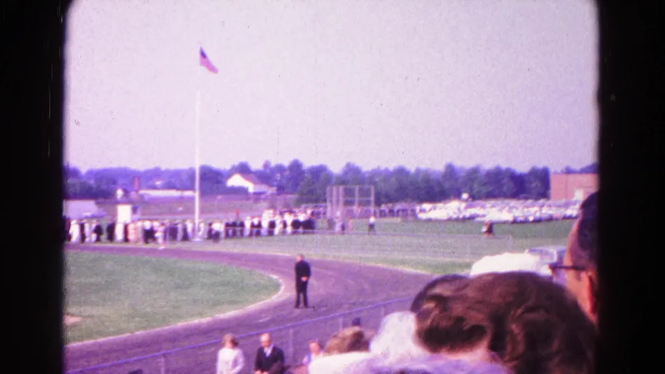 1966 Hagerstown spectator view of a track field ceremony with a long line of people, an American flag, and heads of viewers in the foreground.