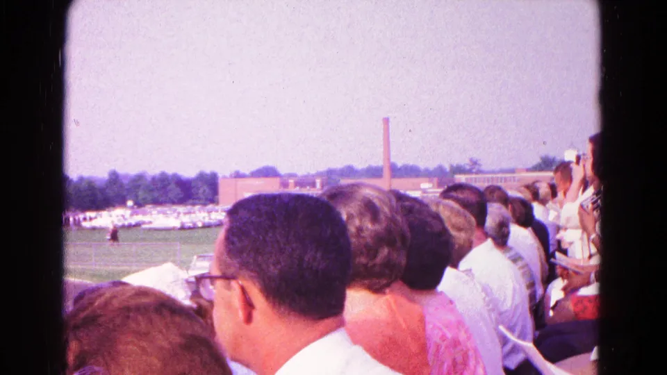 1960s home-movie view from crowded bleachers in Hagerstown, with seated spectators overlooking the school field and ceremony area.