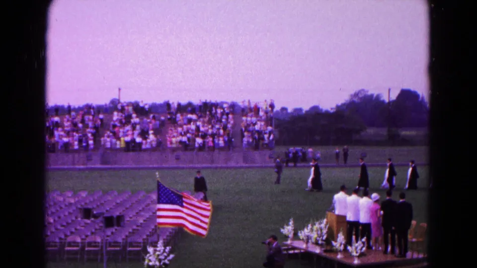 Hagerstown graduation ceremony frame with an American flag, rows of chairs, distant bleachers, and figures in academic gowns on the field.