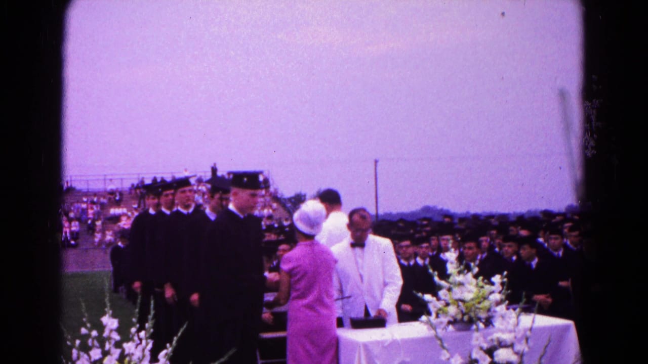 Graduates in caps and gowns approach a diploma table with flowers.