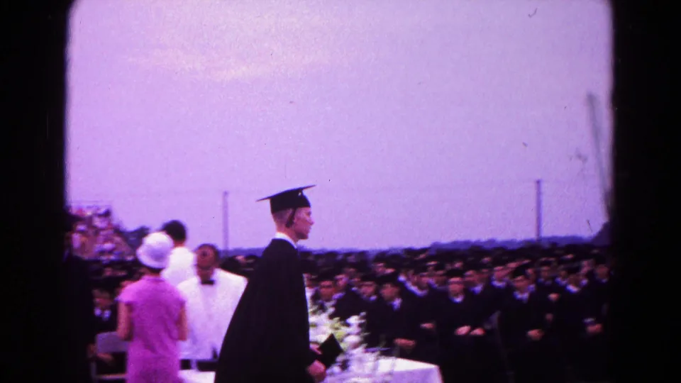 A cap-and-gown graduate passes the ceremony table in Hagerstown, Maryland, with seated graduates and spectators behind.
