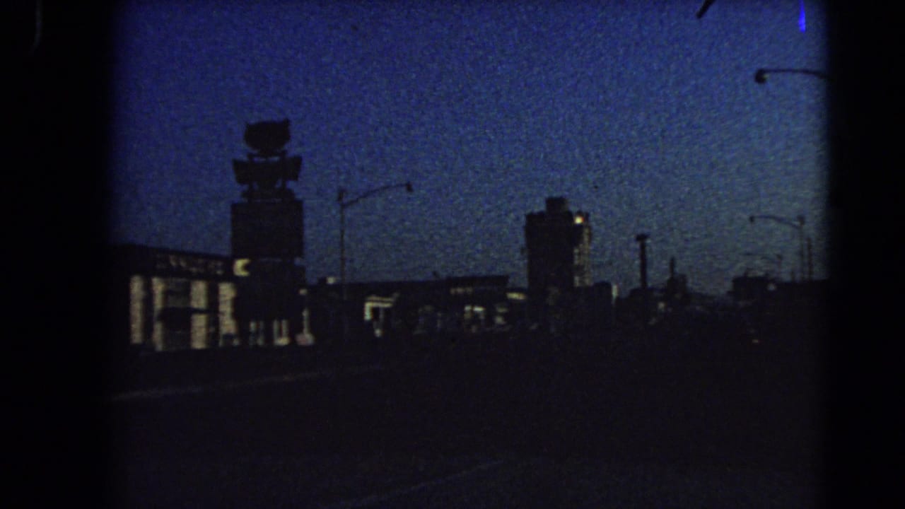 1966 Las Vegas home-movie frame showing a dark roadway and silhouetted buildings beneath a grainy blue dusk sky.