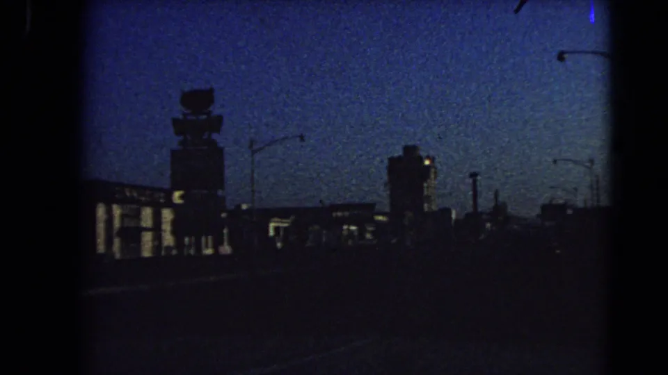 1966 Las Vegas home-movie frame showing a dark roadway and silhouetted buildings beneath a grainy blue dusk sky.