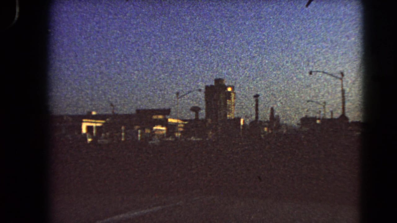 1966 Las Vegas roadway view from a moving vehicle, with silhouetted buildings, streetlights, and a tall central structure at dawn.