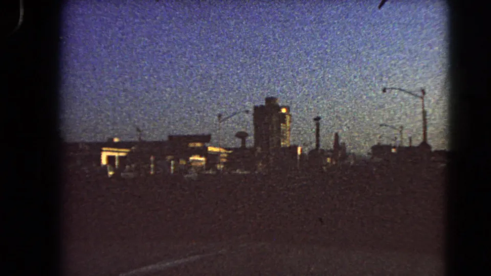 1966 Las Vegas roadway view from a moving vehicle, with silhouetted buildings, streetlights, and a tall central structure at dawn.