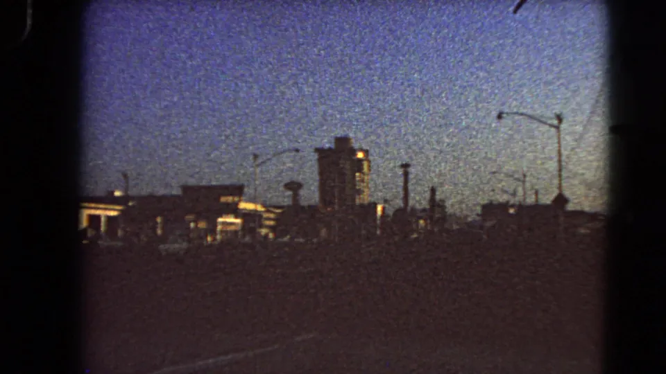 1966 Las Vegas skyline from a vehicle, showing low buildings, streetlights, and a prominent tall building against early morning light.