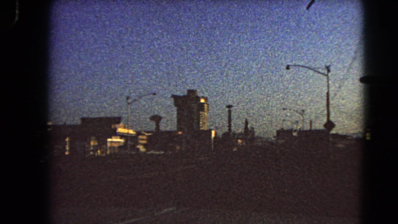 Moving 8mm view of Las Vegas in 1966, with a tall central building, streetlights, and roadway foreground in grainy dawn exposure.