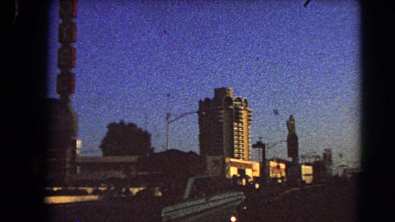 1966 Las Vegas 8mm frame showing a high-rise hotel beyond roadside buildings, with a streetlight, vertical sign, and car at dusk.