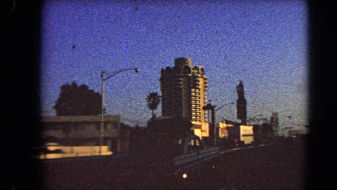 1966 Las Vegas roadside frame with the hotel tower, low foreground structures, a streetlight, and dark 8mm frame borders at dusk.