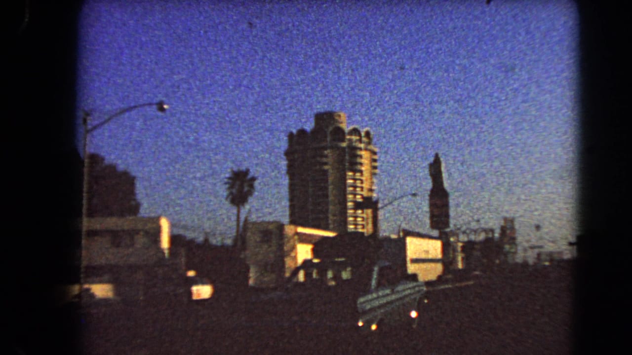 1966 Las Vegas home-movie frame showing a car, streetlight, palm, vertical sign, and high-rise hotel from a moving roadside viewpoint.