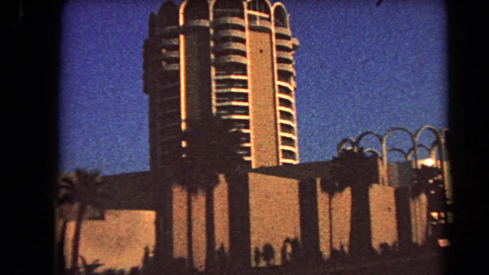 1966 Las Vegas 8mm frame with a closer view of the rounded-balcony high-rise, low foreground wall, palms, and decorative roadside forms.