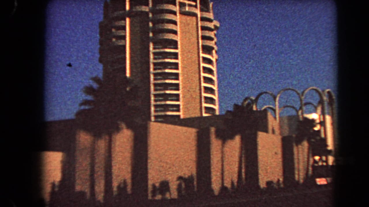 1966 Las Vegas 8mm frame showing a modern high-rise, palms, and arched facade elements against a deep blue dusk sky.