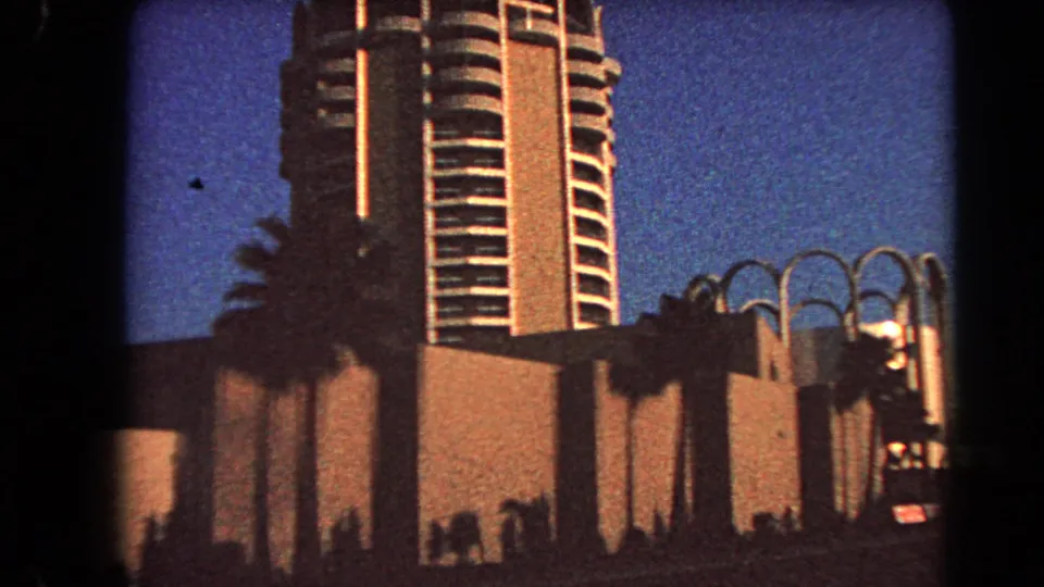 1966 Las Vegas 8mm frame showing a modern high-rise, palms, and arched facade elements against a deep blue dusk sky.