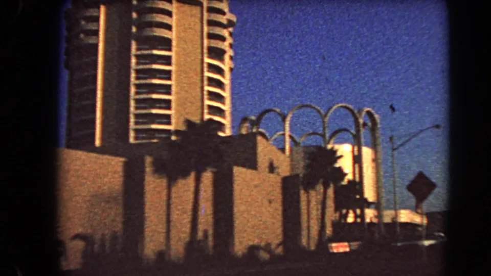 1966 Las Vegas exterior with a tall tower, palm shadows, curved facade details, and a dim street foreground.