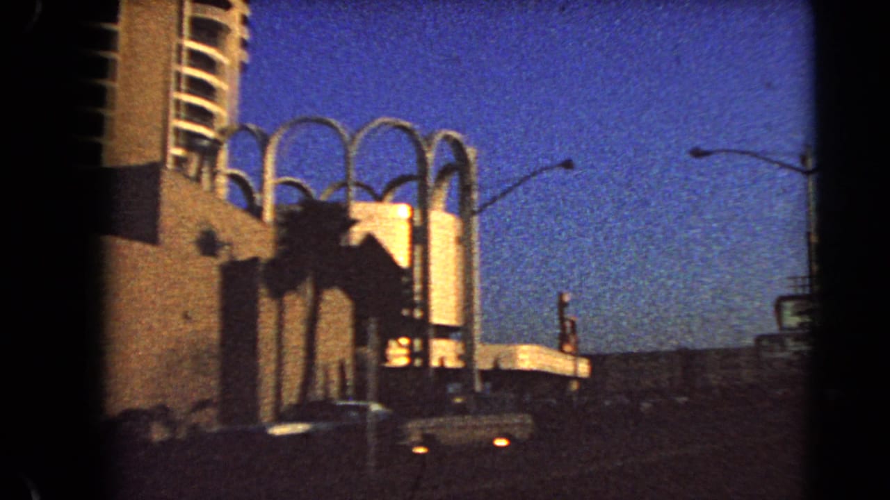 1966 Las Vegas 8mm view of arched architectural features, palms, parked or passing cars, and a streetlight under twilight sky.