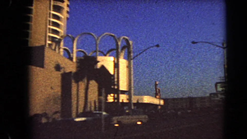 1966 Las Vegas 8mm view of arched architectural features, palms, parked or passing cars, and a streetlight under twilight sky.