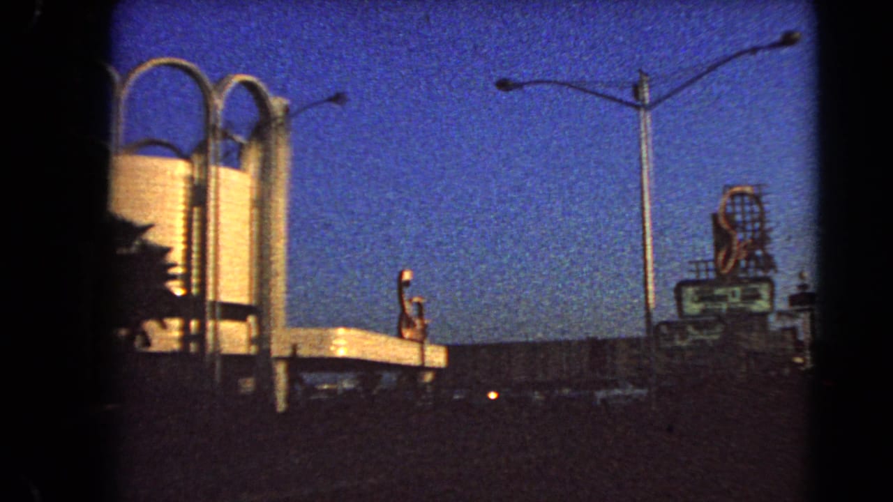 1966 Las Vegas street view with arched building details, a tall streetlight, distant sign shapes, cars, and deep blue evening sky.