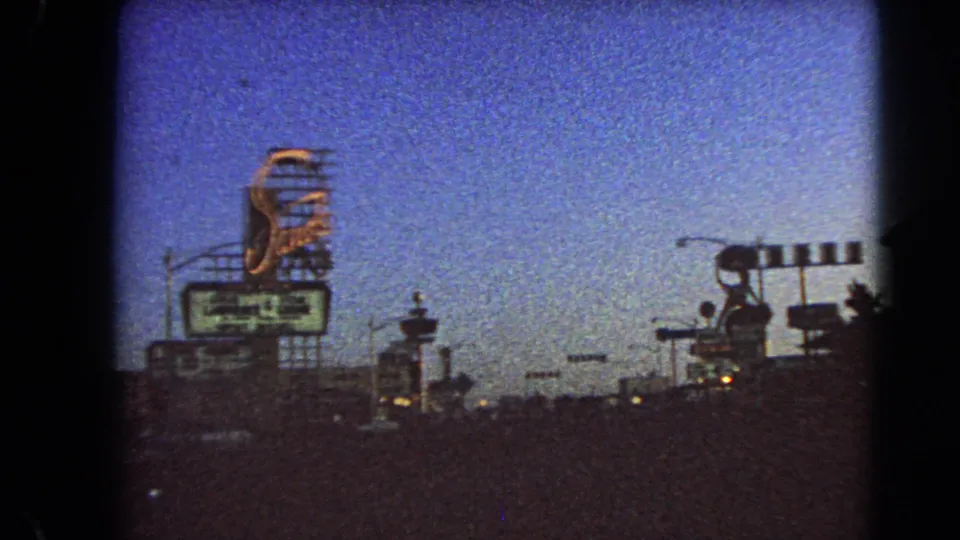 Late-frame 1966 Las Vegas driving view with a large sign, gate-like roadside lights, street poles, and shadowed traffic.