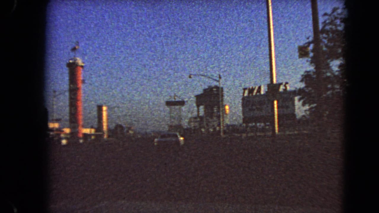 Dusk street view with TWA Jets billboard, Shell sign, red tower, streetlights, and cars.