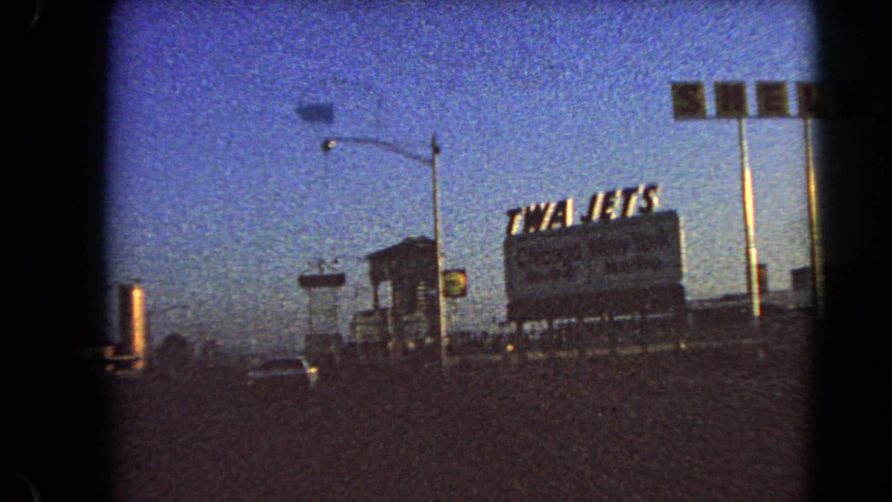 Late clip frame from 1966 Las Vegas showing the TWA Jets billboard, Shell sign, traffic signal, and a car on a dusk street.