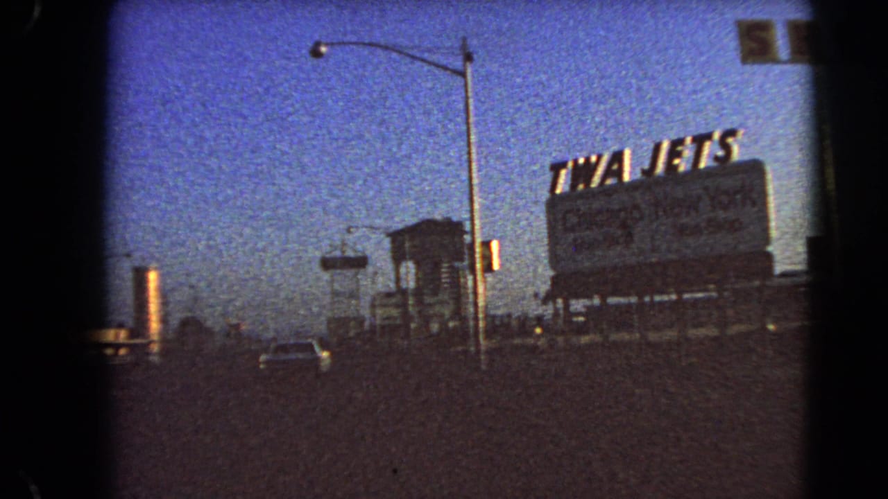Drive-by 8mm frame in Las Vegas showing a large TWA Jets billboard near the road, with streetlights and a deep blue twilight sky.